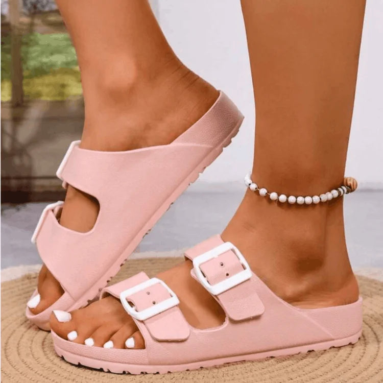 Pink sandals with white buckles worn by a person on a blurred outdoor background