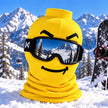 A bright yellow ski face mask with a printed happy face and black goggles, displayed in the snow with snowy mountains and a snowboard in the background.