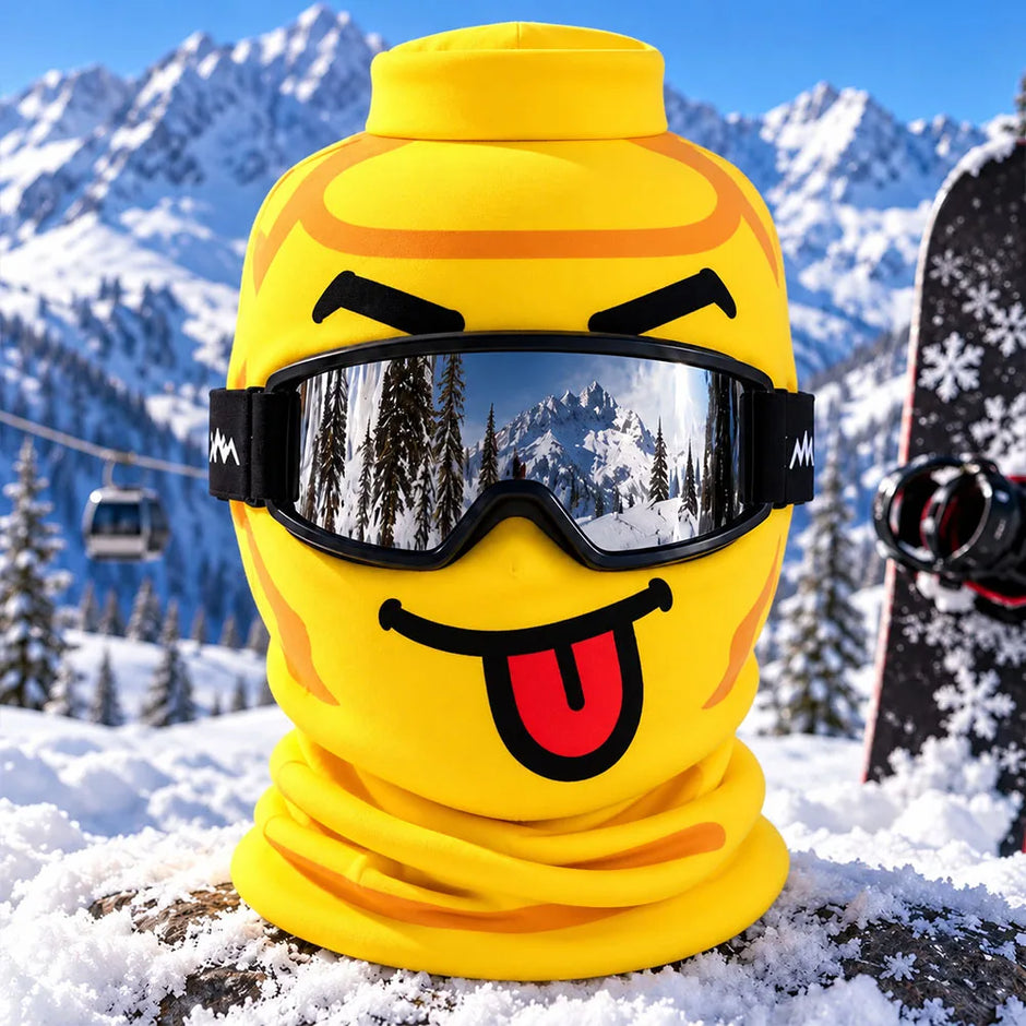 A bright yellow ski face mask with a cartoonish face graphic and black goggles, shown outdoors in a snowy mountain setting with a snowboard and cable cars in the background.