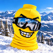 A bright yellow ski mask balaclava with a cartoonish grinning face and black goggles, shown outdoors in the snow with mountains and pine trees in the background.