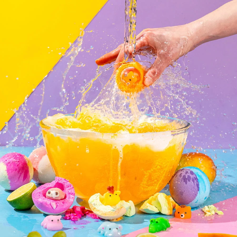 Colorful bath bombs being dropped into a glass bowl filled with water, with a vibrant yellow and purple background.
