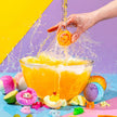 Colorful bath bombs being dropped into a glass bowl filled with water, with a vibrant yellow and purple background.