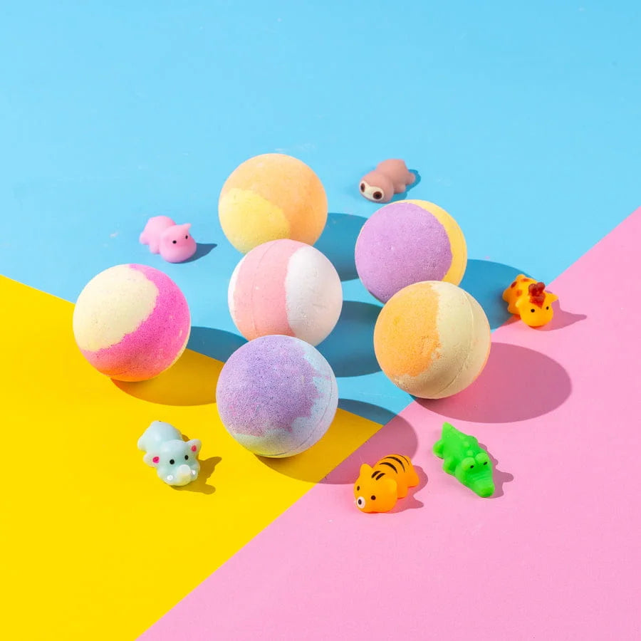 Colorful bath bombs on a pink and blue geometric background with small rubber duck toys.