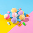Colorful bath bombs on a pink and blue geometric background with small rubber duck toys.