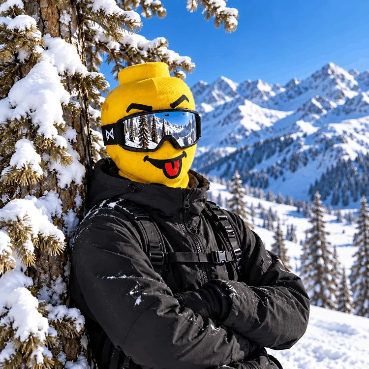 A person in a snowy mountain setting wearing a bright yellow novelty helmet mask with a smiling face, black ski goggles, and a black winter jacket, leaning against a snow-covered tree.