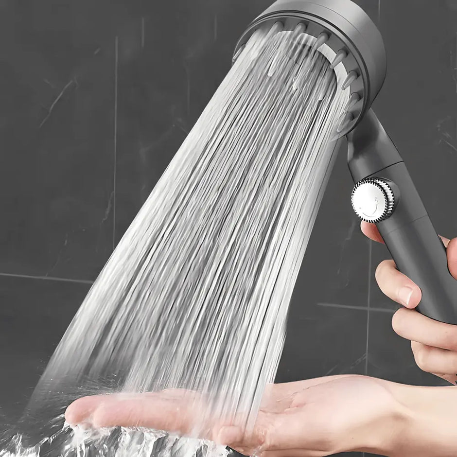 Person holding a shower head with water flowing out, against a dark tiled wall background.