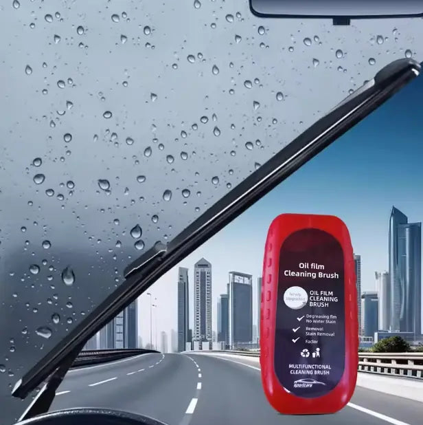 Red oil film cleaning brush held against a car windshield with raindrops and cityscape background