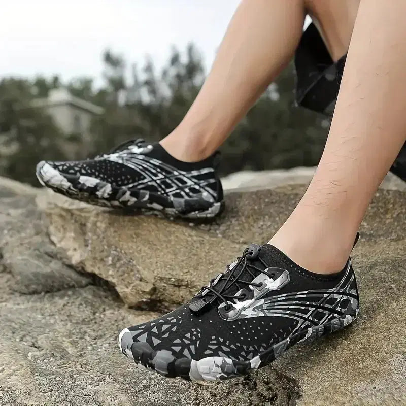 Black and white patterned water shoes worn by a person on rocks with a blurred natural background