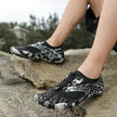 Black and white patterned water shoes worn by a person on rocks with a blurred natural background