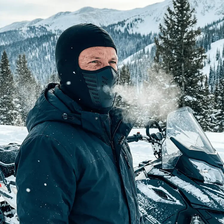 Person in winter gear standing next to a snowmobile in a snowy mountain landscape