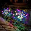 Colorful garden lights in the shape of flowers on a patio with a fence and plants in the background.