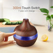 Wooden diffuser with blue LED light on a table with a hand touching the switch, surrounded by plants and fruits.
