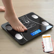 Person using a digital body fat scale with a smartphone app on a wooden floor.